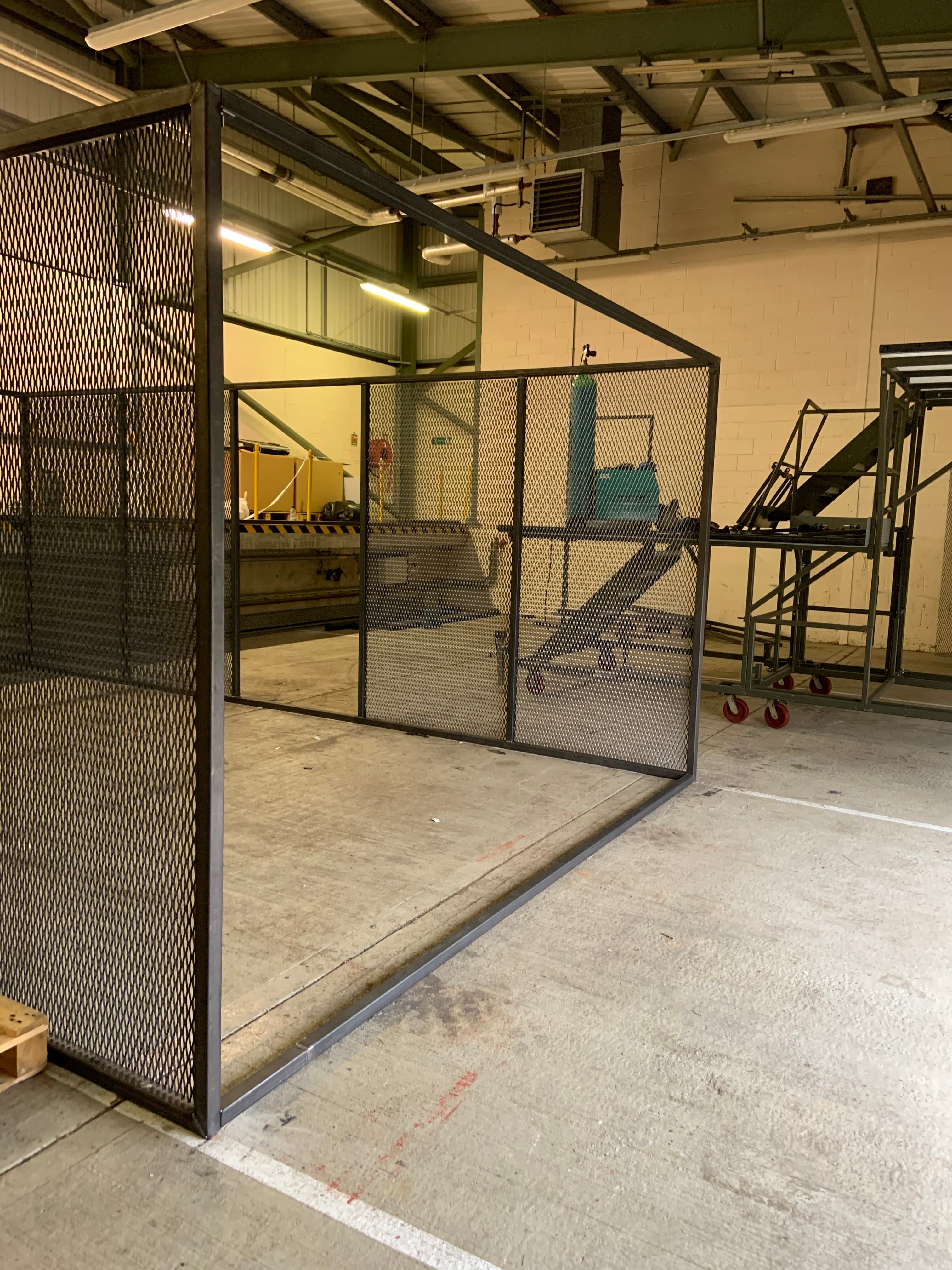Large black metal mesh enclosure in an industrial warehouse with concrete floors and overhead lighting.